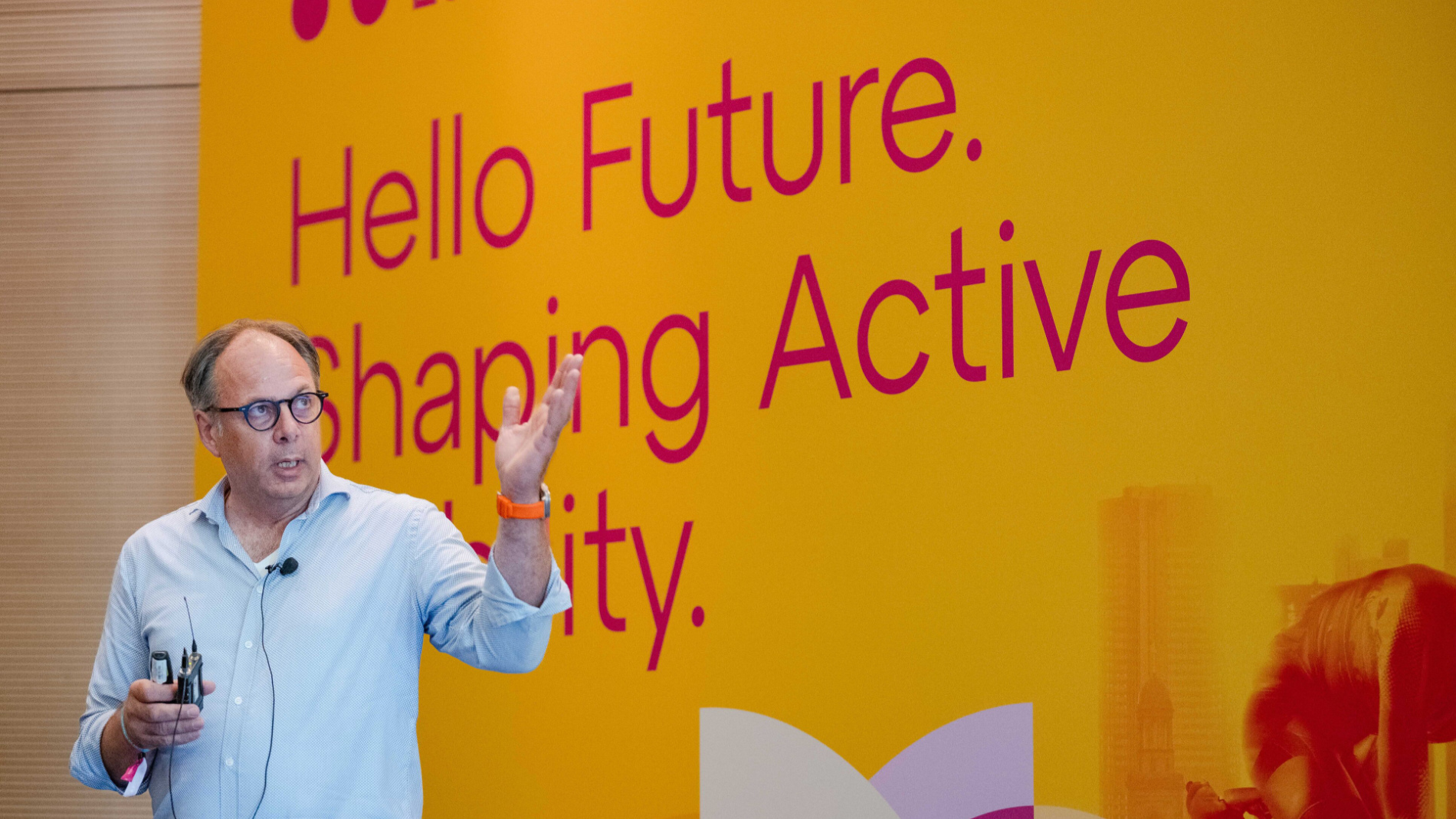 Speaker presenting on stage in front of a yellow backdrop reading “Hello Future. Shaping Active Mobility.”