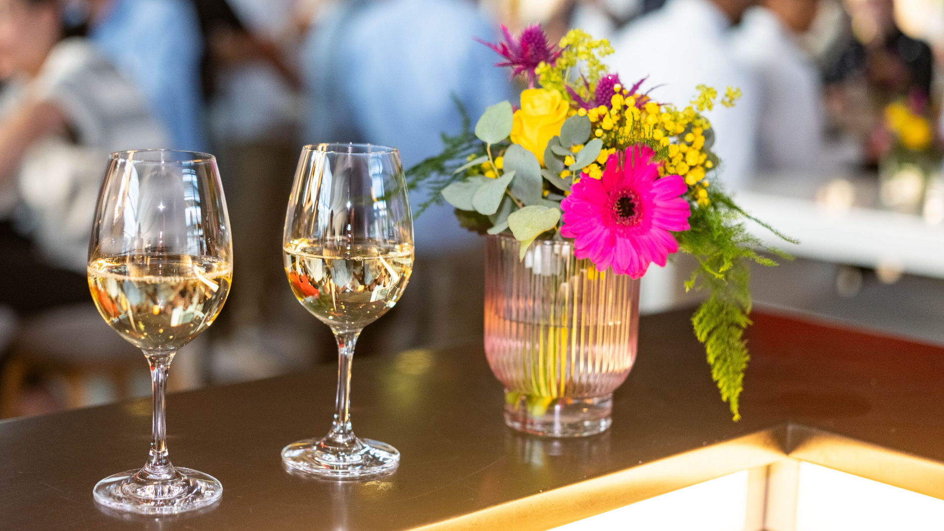 Two glasses of white wine next to a colorful flower arrangement on a bar during an event.